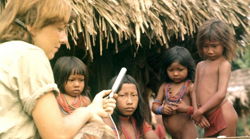 a woman crouched at a tape recorder with several small children in front of a thatched hut