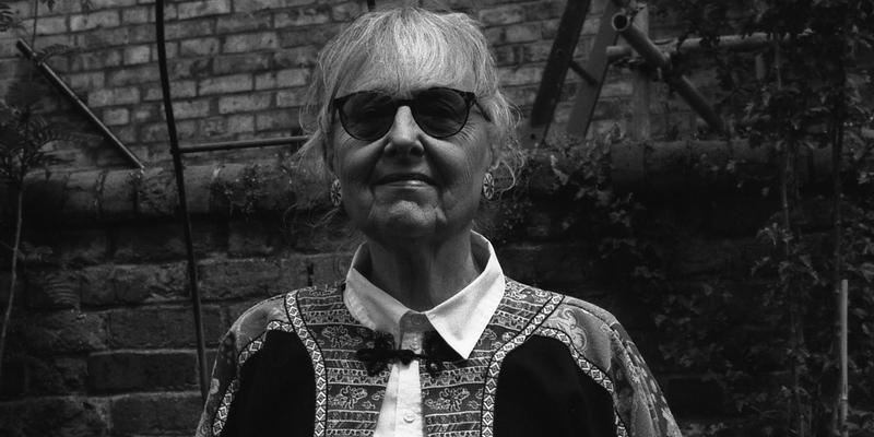 A woman in her 80th year wearing sunglasses, looking defiant and smiling slightly, black and white