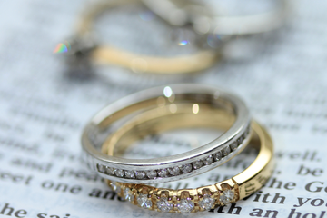 A silver diamond ring stacked on top of a gold diamond ring, resting on a page. In the background are two other rings.
