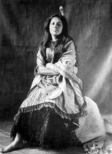 A black and white photo of Mākereti Papakura. She is sitting down with her arms folded, wearing a garment of loose, light-coloured fabric over a dark skirt.