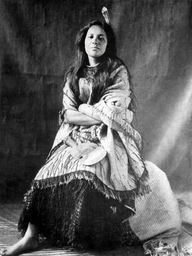 A black and white photo of Mākereti Papakura. She is sitting down with her arms folded, wearing a garment of loose, light-coloured fabric over a dark skirt.