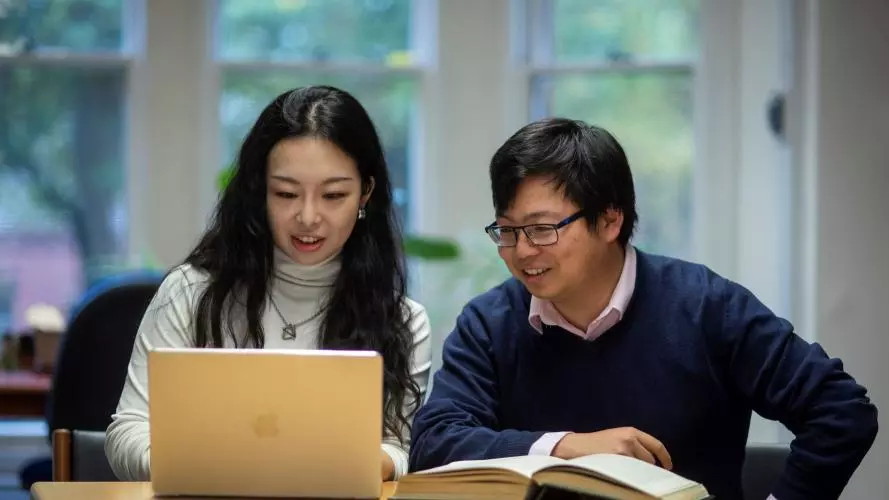 Two students sat together at a desk with a book and a laptop.