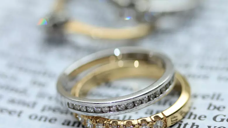 A silver diamond ring stacked on top of a gold diamond ring, resting on a page. In the background are two other rings.