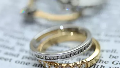 A silver diamond ring stacked on top of a gold diamond ring, resting on a page. In the background are two other rings.