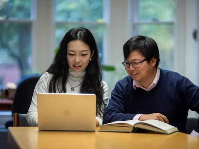 Two students sat together at a desk with a book and a laptop.