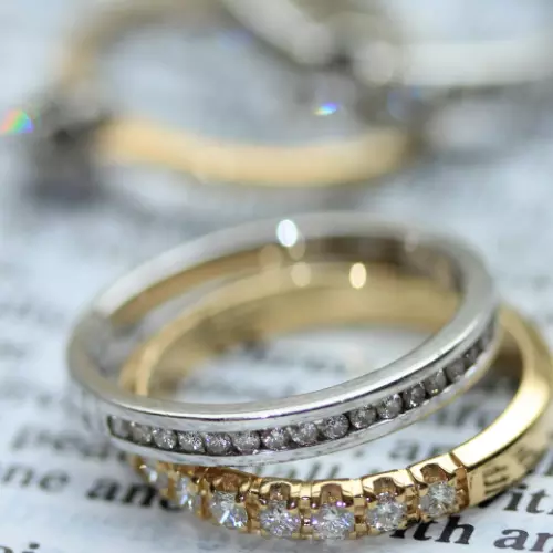 A silver diamond ring stacked on top of a gold diamond ring, resting on a page. In the background are two other rings.