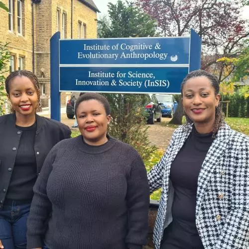 Three women standing in a line outside the department buildings, under a sign for the Institute for Science, Innovation and Society (InSIS)