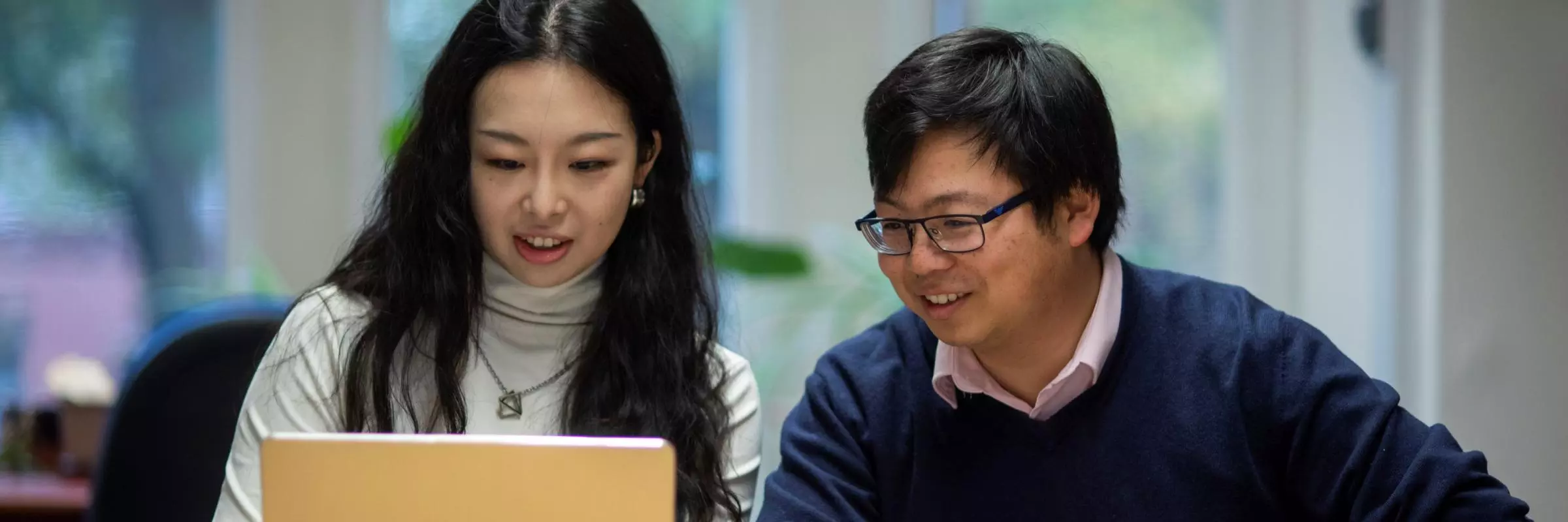 Two students sat together at a desk with a book and a laptop.
